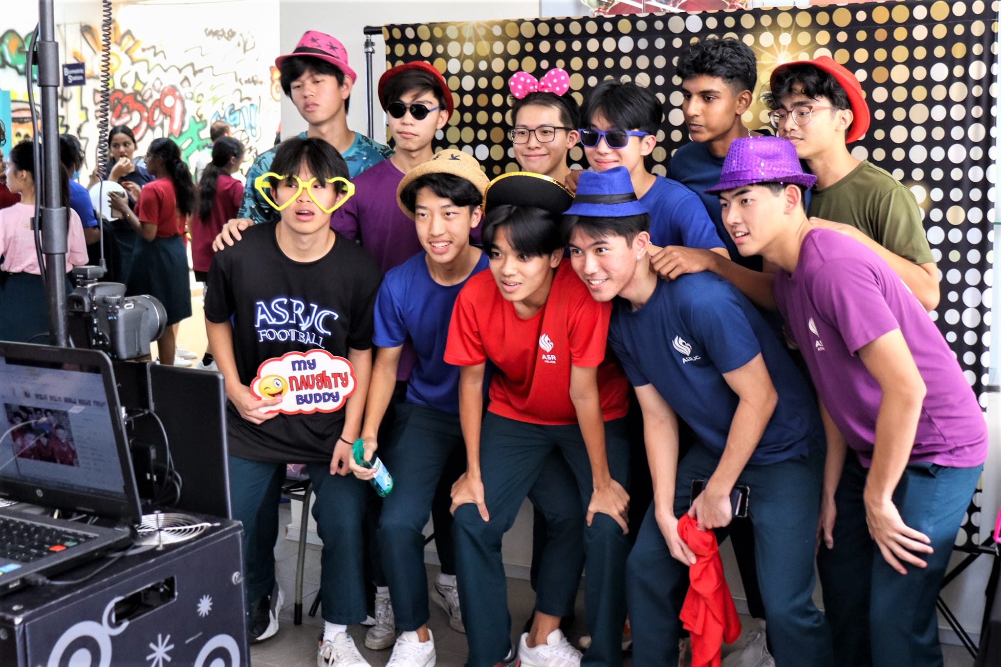 A group of teens wearing colorful hats and props pose together at a photo booth with a polka dot backdrop.
